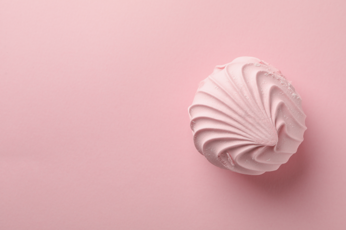 Une délicate meringue rose pastel, avec une texture lisse et satinée, posée sur un fond rose uni, symbolisant une préparation culinaire douce et aérienne aromatisée aux huiles essentielles.