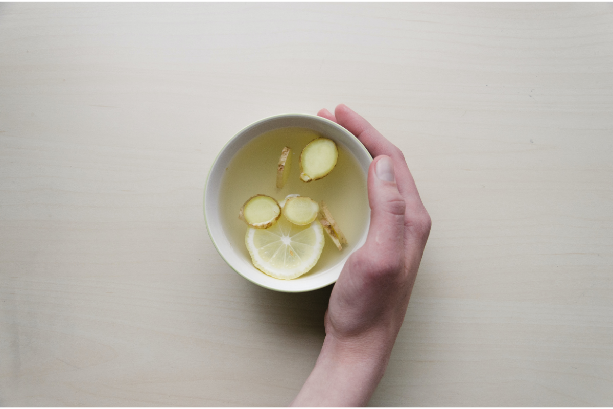 Une main tenant une tasse blanche contenant une boisson chaude claire avec des tranches de gingembre et de citron flottant à la surface, posée sur une table au ton bois clair.