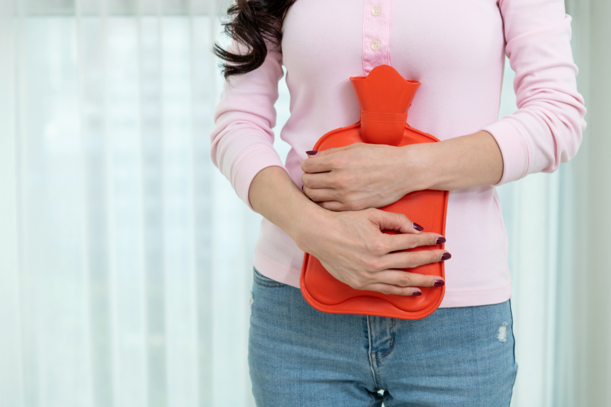 Une femme tenant une bouillotte en caoutchouc rouge contre son abdomen pour soulager la douleur. Image illustrant l'usage thérapeutique de la bouillotte en naturopathie, notamment pour apaiser les douleurs menstruelles et favoriser la détente.