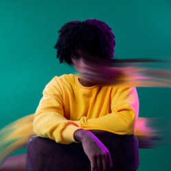Portrait d'une personne assise, vêtue d'un pull jaune vif, avec une traînée floue de couleurs qui traverse l'image, évoquant un sentiment de mouvement ou de tourbillon d'anxiété, sur un fond vert uni.