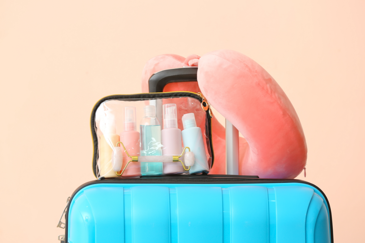 Une valise bleue équipée d'une trousse de toilette transparente contenant des flacons de voyage aux couleurs pastel, symbolisant la préparation d'une trousse de secours pour les vacances. Un coussin de voyage rose est posé sur le manche de la valise, évoquant le confort et la mobilité.
