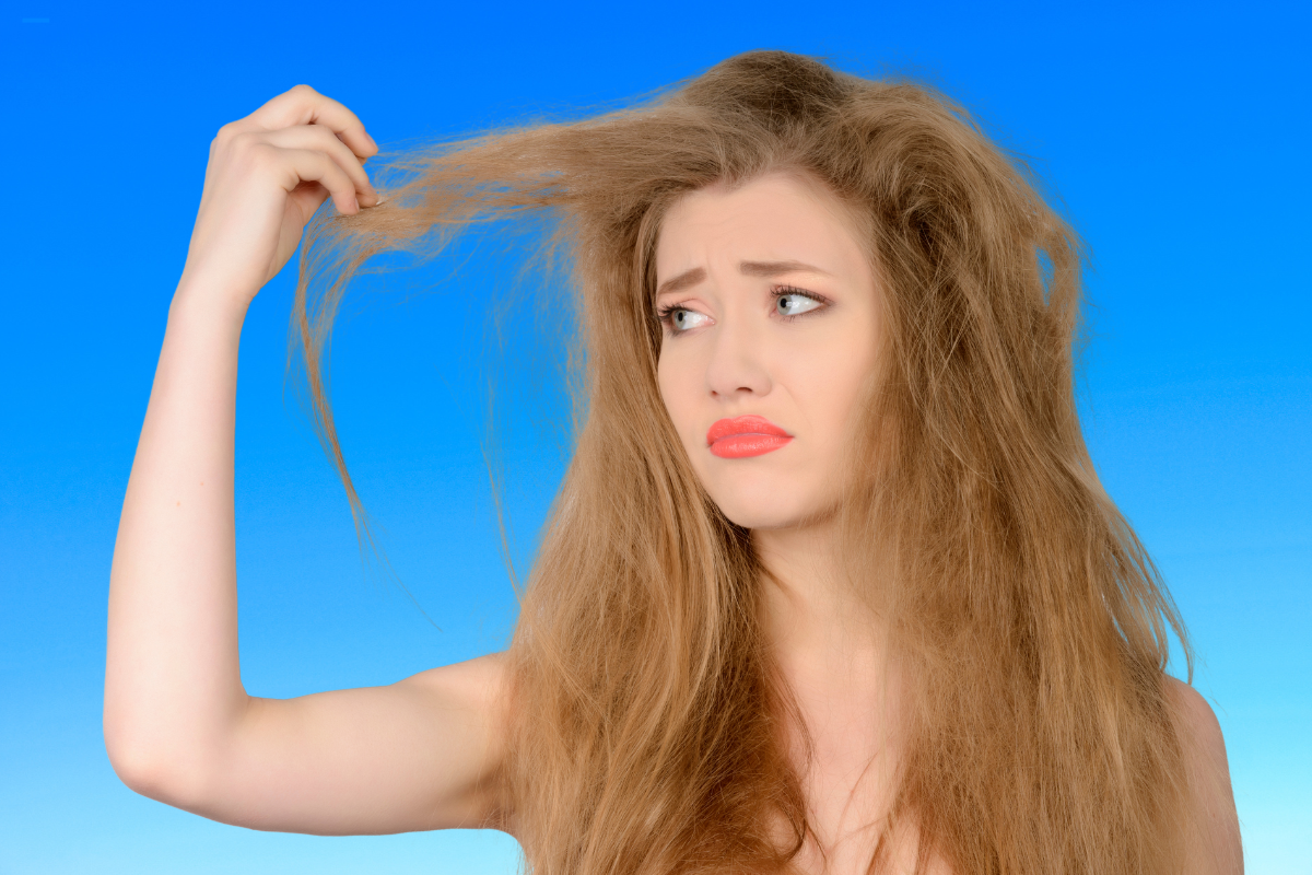 Una mujer con el cabello seco y dañado tira de un mechón, ilustrando los efectos de la sequedad capilar y la necesidad de cuidados nutritivos a base de aceites vegetales.