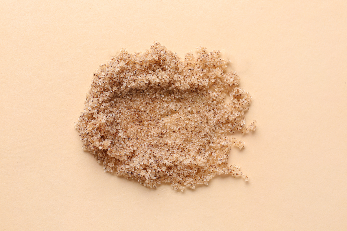 Une petite quantité de gommage pour le corps à base de sucre brun repose sur une surface de couleur beige claire. La texture granuleuse et irrégulière du gommage est clairement visible, indiquant sa nature exfoliante. La couleur dorée du sucre brun offre un contraste subtil avec l'arrière-plan neutre.