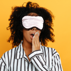 Donna in pigiama con maschera per il sonno rosa che sbadiglia davanti a uno sfondo giallo, a simboleggiare i disturbi del sonno e le soluzioni naturali come i Fiori di Bach.