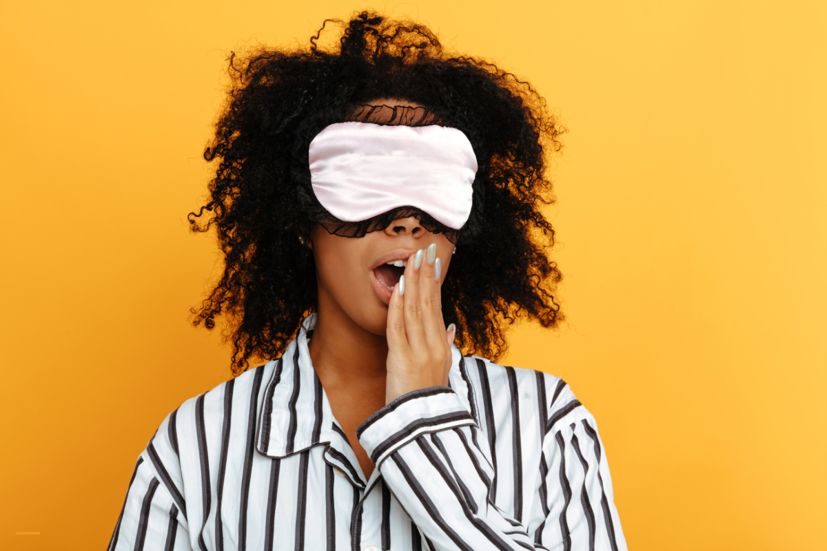 Vrouw in pyjama met een roze slaapmasker geeuwt voor een gele achtergrond, symbool voor slaapstoornissen en natuurlijke oplossingen zoals Bach bloesemremedies.