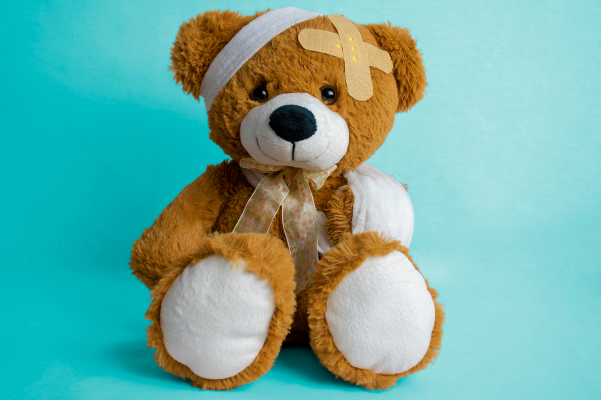 Ours en peluche marron avec un bandage sur la tête et des pansements adhésifs sur le corps posé contre un fond bleu uni.