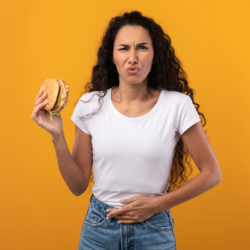 Uma mulher com um rosto perturbado, segurando um hambúrguer numa mão e o estômago na outra, expressando desconforto digestivo, num fundo amarelo vivo.