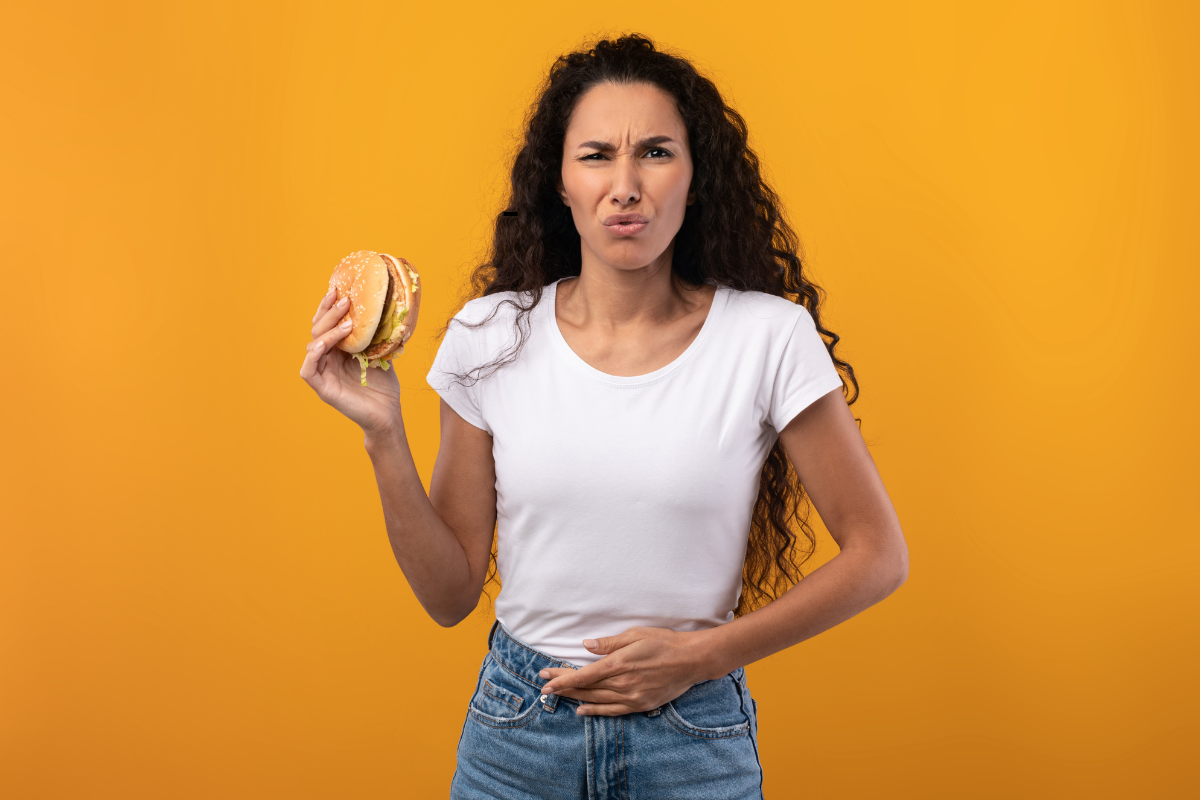 Een vrouw met een overstuur gezicht die een hamburger in haar ene hand houdt en haar maag in de andere, om spijsverteringsproblemen uit te drukken, tegen een felgele achtergrond.