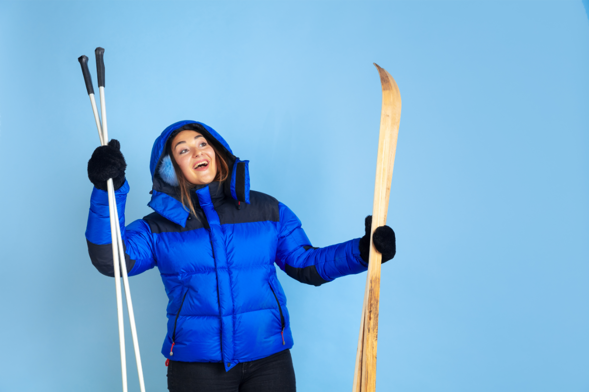Une femme souriante en tenue d'hiver bleue tenant des skis et des bâtons de ski se prépare à profiter des sports d'hiver.