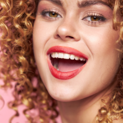 Une femme souriante avec des cheveux bouclés et un rouge à lèvres rouge vif affiche un sourire large et éclatant qui met en évidence des dents blanches et bien entretenues, symbolisant la joie et la santé dentaire.