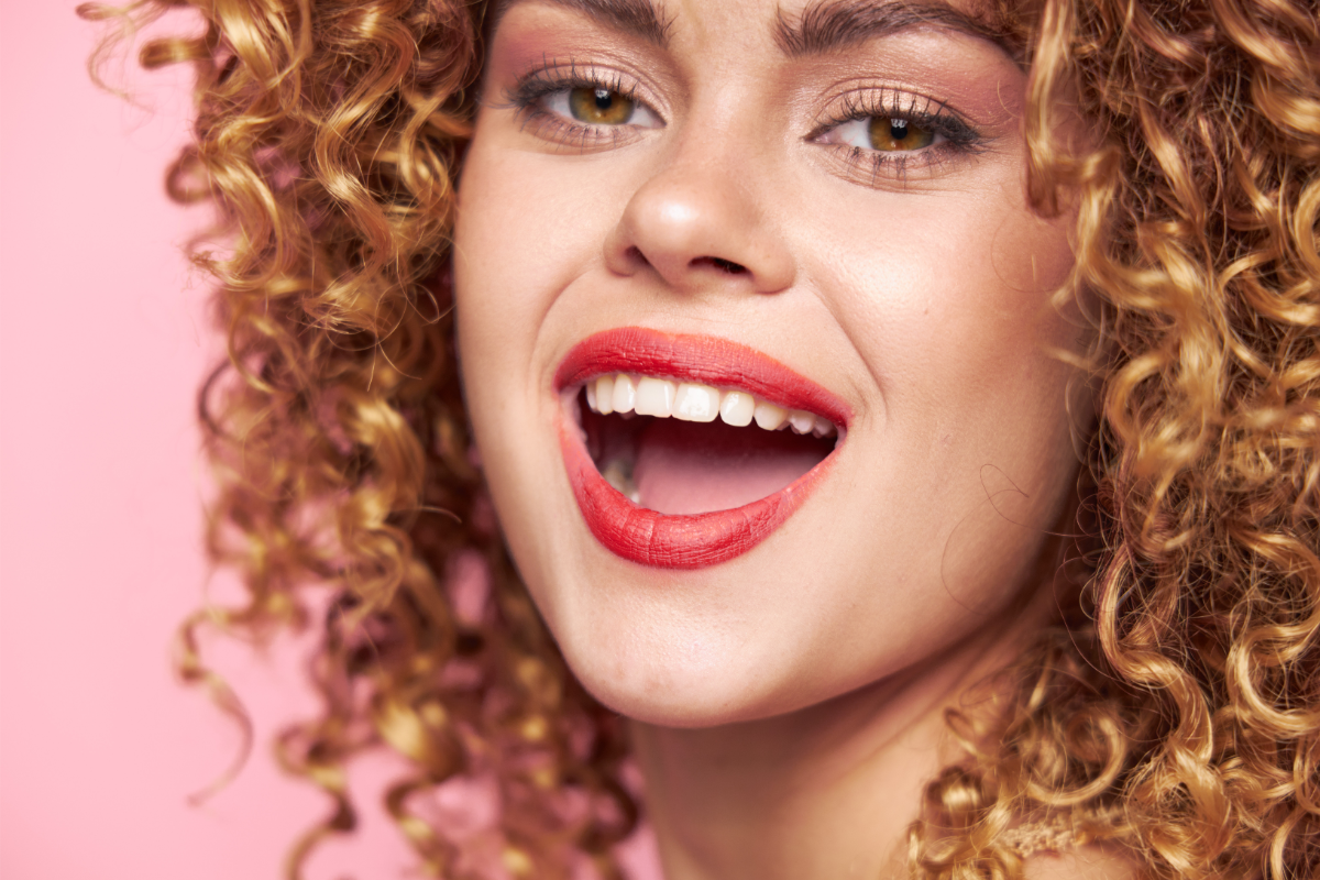 Une femme souriante avec des cheveux bouclés et un rouge à lèvres rouge vif affiche un sourire large et éclatant qui met en évidence des dents blanches et bien entretenues, symbolisant la joie et la santé dentaire.
