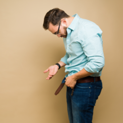Homme en chemise bleue à carreaux et pantalon en jean, se penchant vers l'avant, regardant et pointant vers sa ceinture défaite, avec une expression de déception ou de confusion.