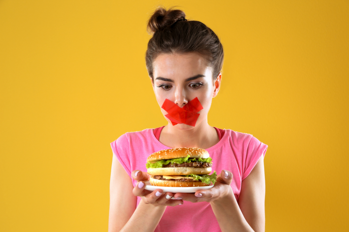 L'image représente une personne avec du scotch sur la bouche. elle tient un hamburger dans ses mains pour illustrer les aliments non autorisés dans le régime AIP. Le régime Paléo A.I.P dans la gestion des maladies auto-immunes