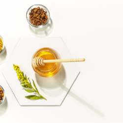 Un pot de miel doré avec une cuillère en bois, accompagné de deux récipients en verre contenant des granules de pollen d'abeille et des morceaux de propolis, avec des fleurs jaunes et des feuilles vertes en décoration, le tout sur un fond blanc épuré.