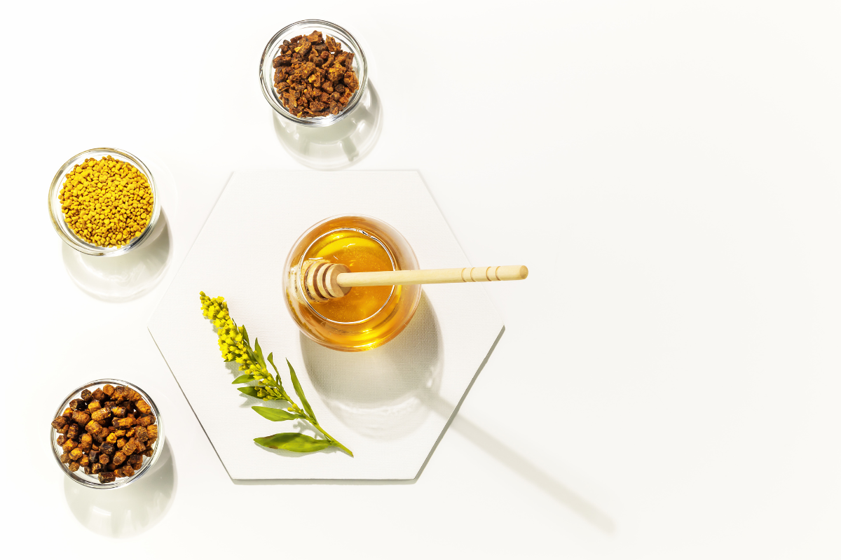 Un pot de miel doré avec une cuillère en bois, accompagné de deux récipients en verre contenant des granules de pollen d'abeille et des morceaux de propolis, avec des fleurs jaunes et des feuilles vertes en décoration, le tout sur un fond blanc épuré.
