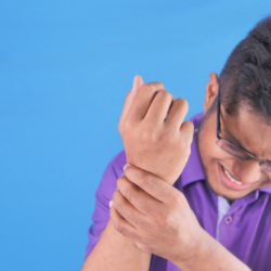 Un homme semble éprouver une vive douleur au poignet gauche, qu'il tient fermement avec sa main droite. Il porte des lunettes et une chemise mauve, et grimace de douleur. Le fond de l'image est uni et bleu, mettant en évidence la réaction de l'homme à la douleur. Cela pourrait illustrer un article sur la gestion de la douleur, notamment par des méthodes comme l'homéopathie.