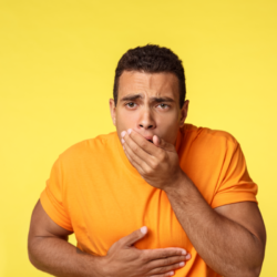 Homme jeune portant un t-shirt orange, tenant sa main sur son ventre et l'autre main sur sa bouche avec une expression faciale indiquant qu'il se sent mal ou nauséeux, sur un fond jaune uni.