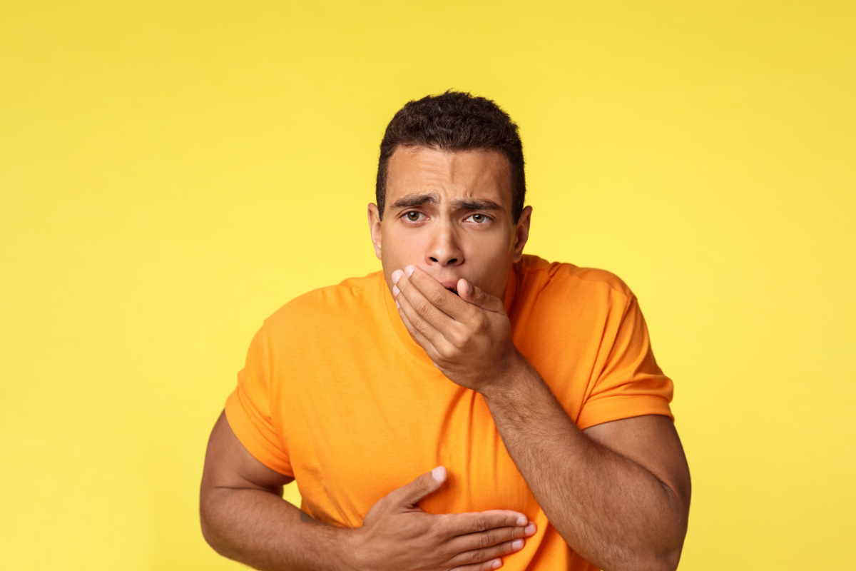 Homme jeune portant un t-shirt orange, tenant sa main sur son ventre et l'autre main sur sa bouche avec une expression faciale indiquant qu'il se sent mal ou nauséeux, sur un fond jaune uni.