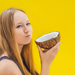 Les bienfaits du oil pulling./ Une jeune femme qui tient une noix de coco, sur fond jaune. elle est en train de pratiquer le oil pulling