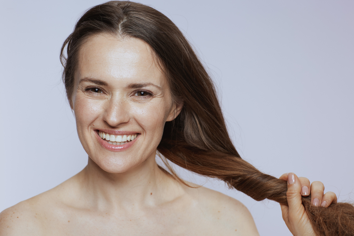 Femme souriante aux cheveux longs et brillants, tenant une mèche entre ses doigts, illustrant les bienfaits des huiles végétales pour des cheveux normaux en bonne santé.