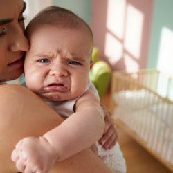 Bebé a chorar nos braços da mãe, demonstrando desconforto ou dor de barriga, num quarto bem iluminado com um berço com grades ao fundo.