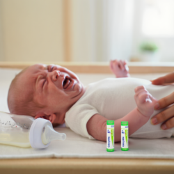 Baby lying down and crying, with a bottle and two homeopathic tubes nearby, illustrating natural remedies for infant colic.