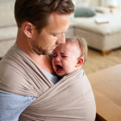 Een vader draagt en wiegt een huilende baby om hem te kalmeren tijdens een koliekaanval, met een flesje op de tafel ernaast.